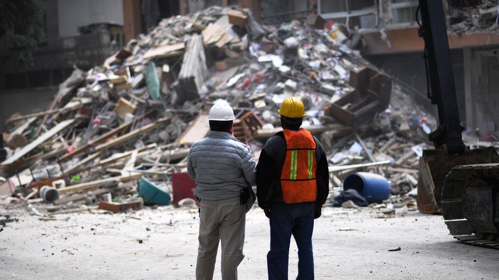 Two men with their backs turned to camera survey a pile of rubble