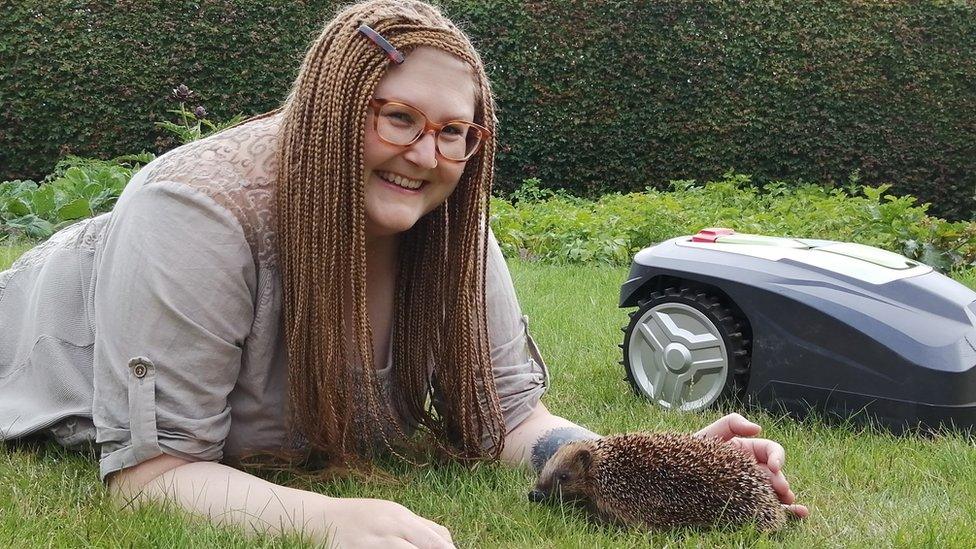 Dr Sophie Lund Rasmussen laying on the grass with a hedgehog next to a robot mower