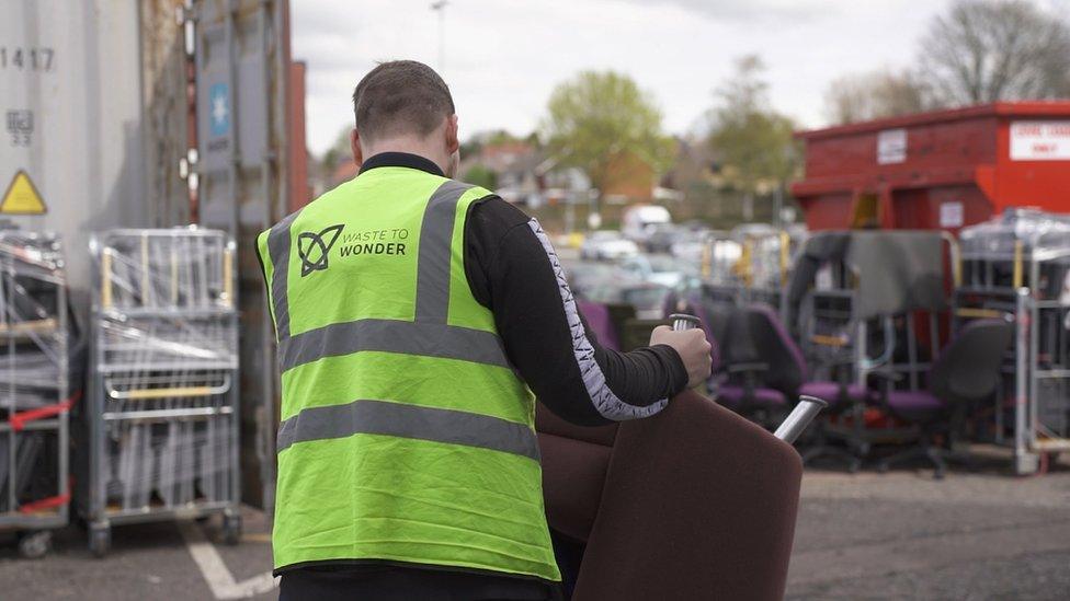 The back of a man in a fluorescent safety jacket holding a chair.