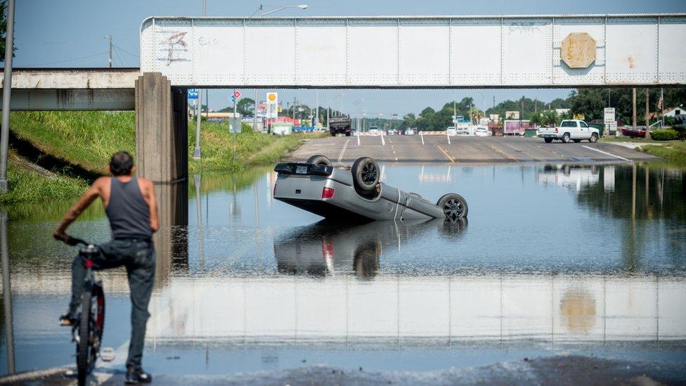 A man on a bicycle surveys an overturned truck in receding floodwater in Port Arthur, Texas