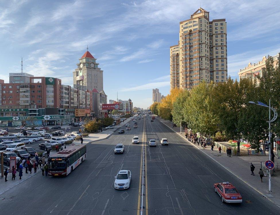 City landscape with cars travelling on a straight road lined by buildings and a car park