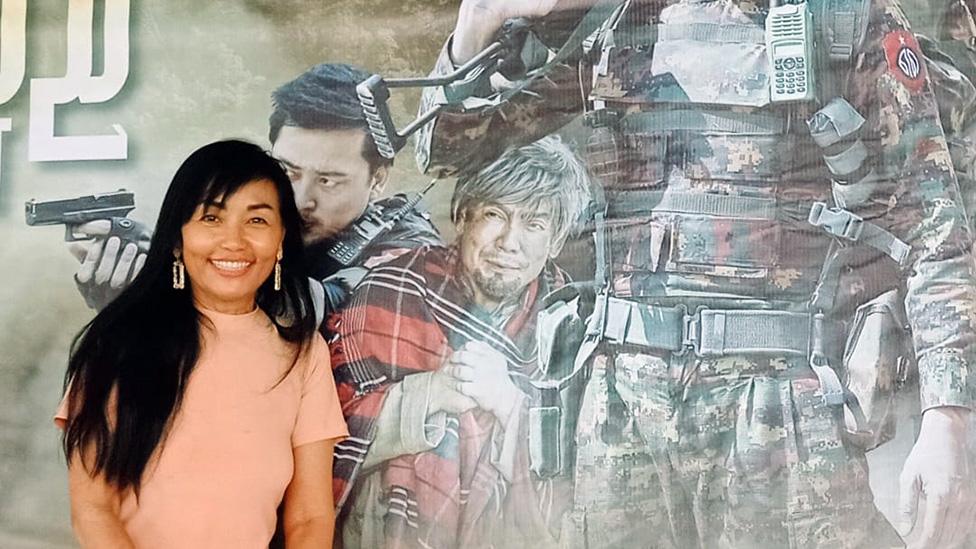 Lily Naing Kyaw posing in front of a poster of a military propaganda film