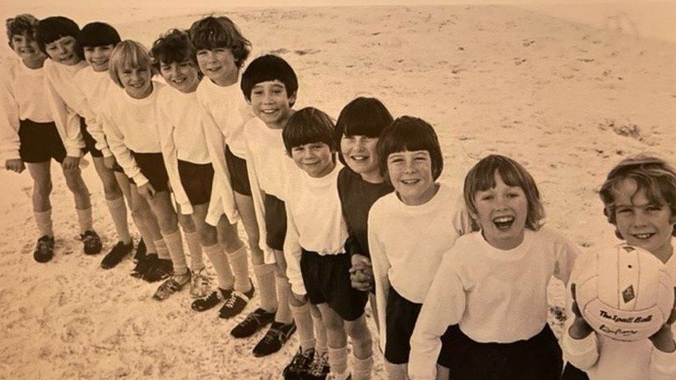 Team of young boys wearing white tops and black shorts. One has a football