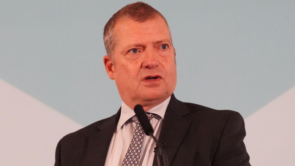 Graham Simpson, who has short grey hair, speaks into a microphone in front of a blue and white sign. He is wearing a black suit, grey shirt and patterned tie.