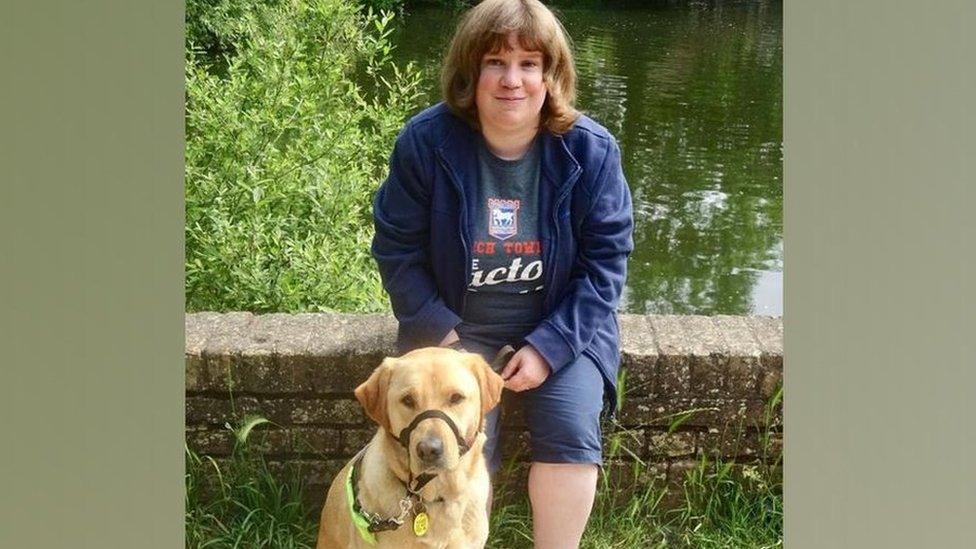 Lisa Newson, dressed in navy blue cropped trousers, an Ipswich Town t-shirt and navy blue jacket sits on a small brick wall. In front of her is her yellow coloured guide dog Holly.