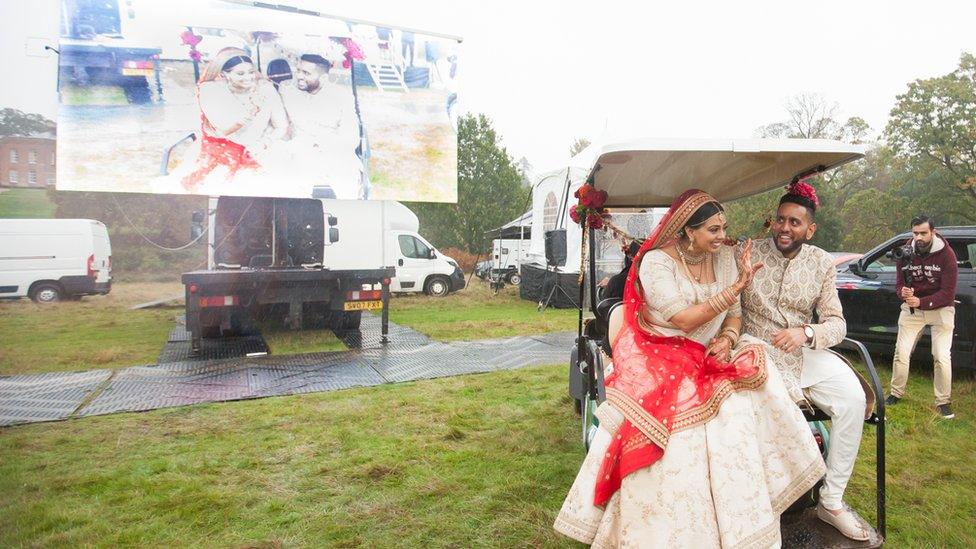 Roma Popat and Vinal Patel on the back of a golf buggy in front of a large screen.