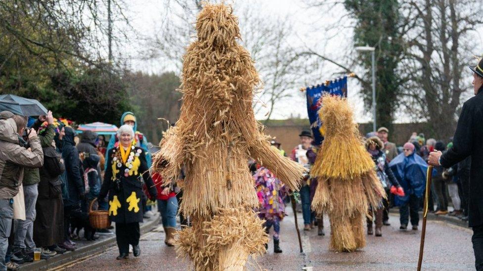 Two figures completely covered with straw walks streets with crowd of people