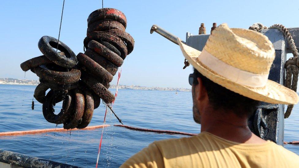 man in straw hat watching tyres being winched out of the sea by a boat