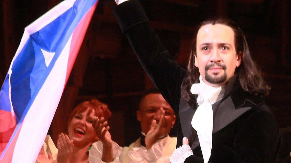 Lin-Manuel Miranda standing on-stage at the end of a performance of Hamilton, holding the Puerto Rico flag