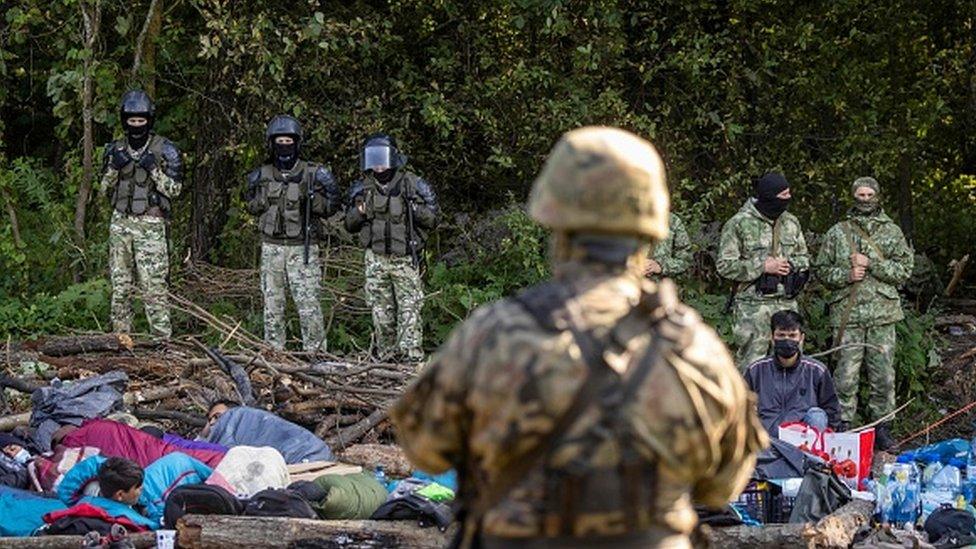 Polish (front) and Belarusian border guards (background) stand next to a group of migrants