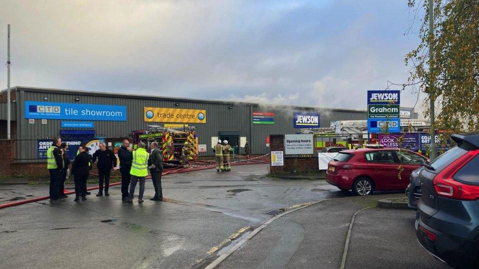 Image of the Jewson warehouse on Woodrow Way in Gloucester. Firefighters and a fire engine can be seen near the entrance of the warehouse and smoke can be seen coming from the building.