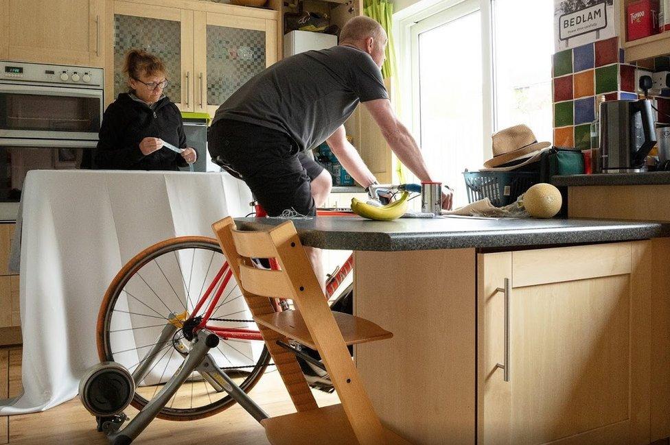 Man cycles on bike inside while wife does chores