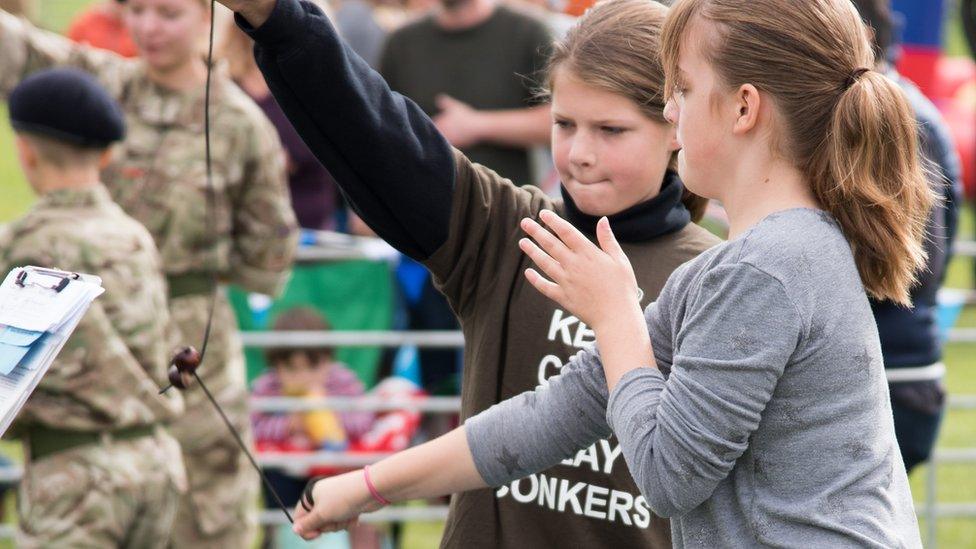 Two girls smash conkers together as someone unseen observes holding a clipboard.