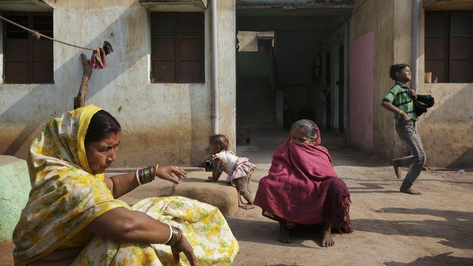 Two Indian women sat opposite each other on the pavement in an Indian village. A boy runs behind them in the background.