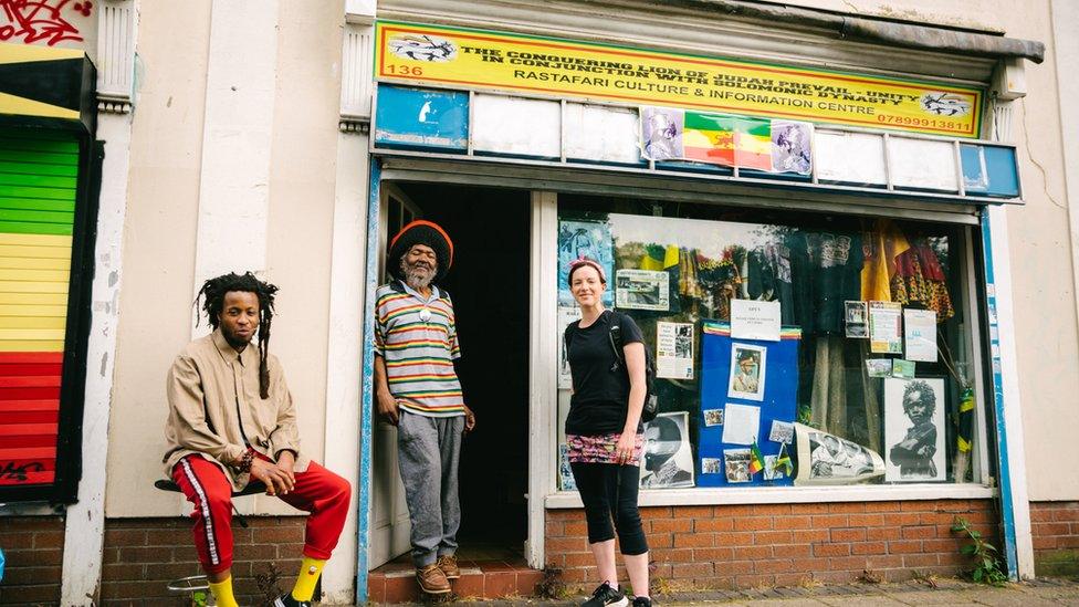 Two people standing and one sitting in front of the Rastafarian Centre, which has a yellow sign