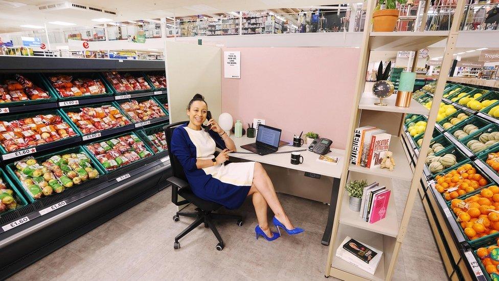 Woman sits at desk in the middle of a fruit and veg supermarket aisle