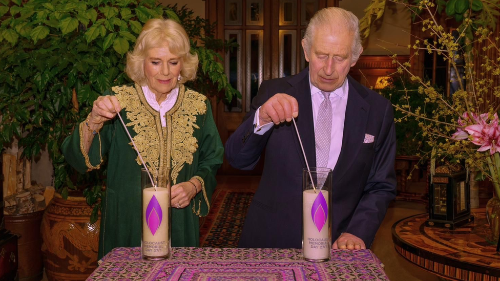 Camilla and Charles stand behind a table on which two large candles decorated with a purple flame. They each hold a long match to the wick.