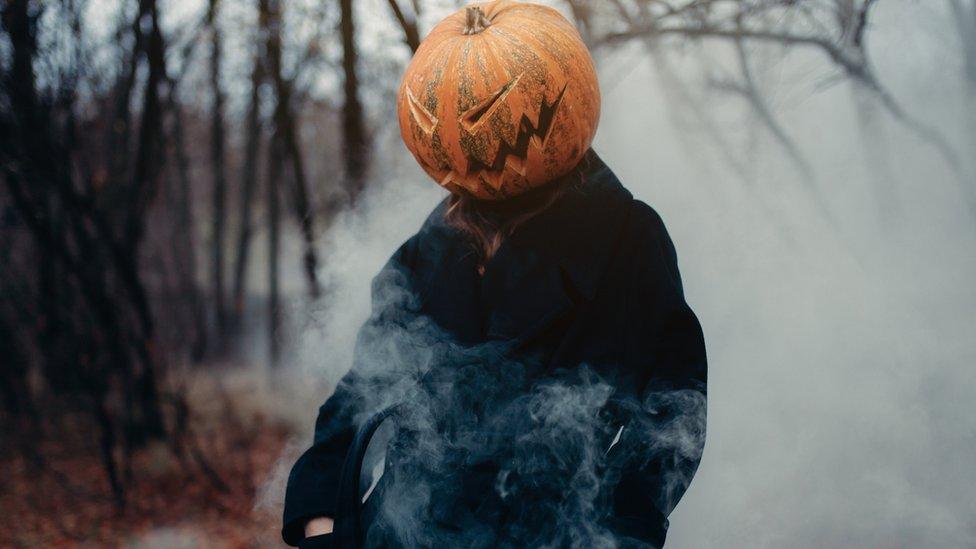 A man in a long black coat with a pumpkin instead of a head in a dark, damp and foggy forest.