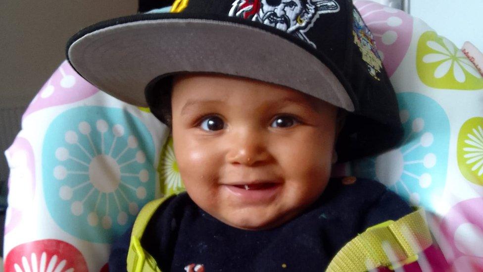 Baby Adonis wearing a hat and shaking a rattle in a baby chair.