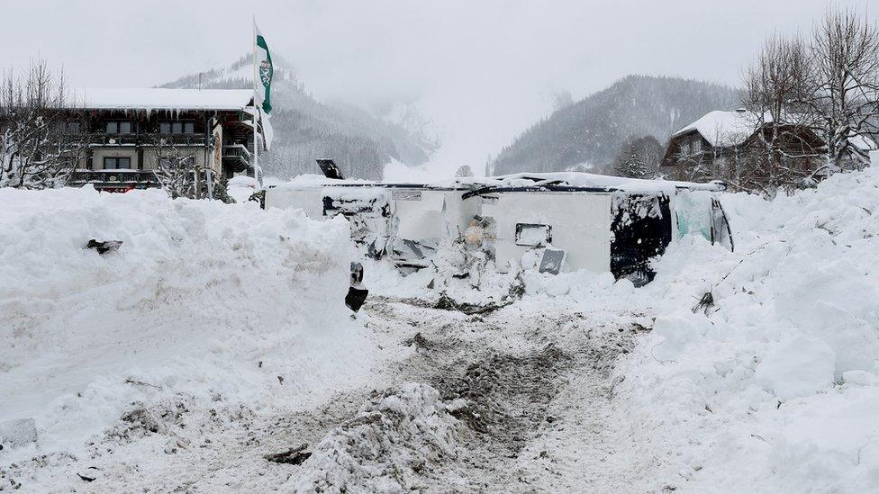 A bus, lying on its side, is seen surrounded by banks of snow equal to its huge size, amid the snowy ski resort town