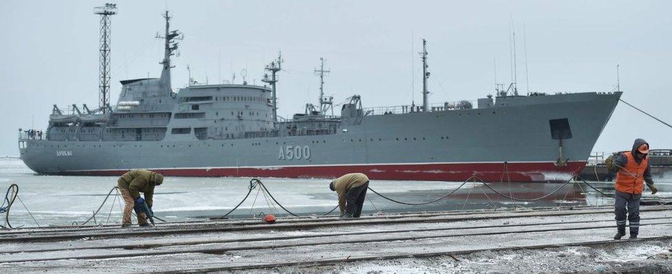 A very large military ship is seen floating on an icy bay in the background, while closer to the camera, a handful of men work on a long, empty pier area