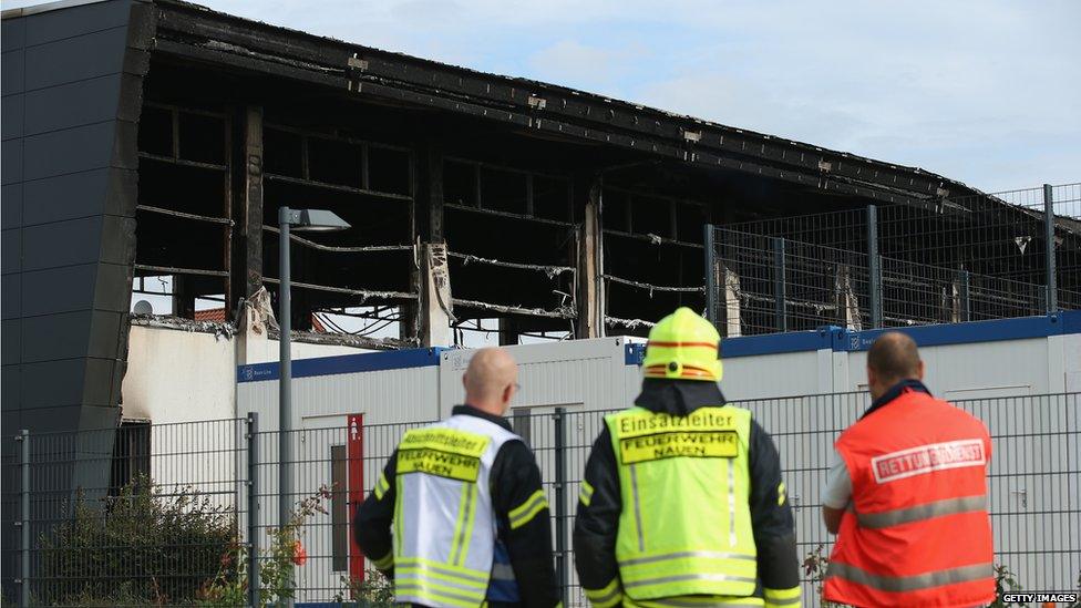 Firemen and an emergency services worker stand outside a sports hall struck by fire that was intended to house refugees and migrants applying for asylum in Germany on 25 August 2015 in Nauen