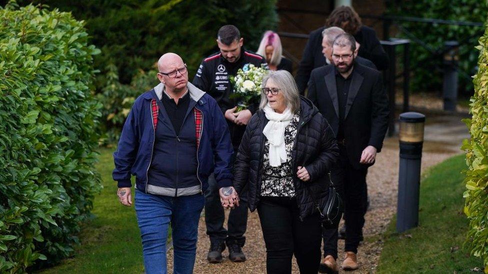 A group of people attending the funeral walk down a tree-lined path