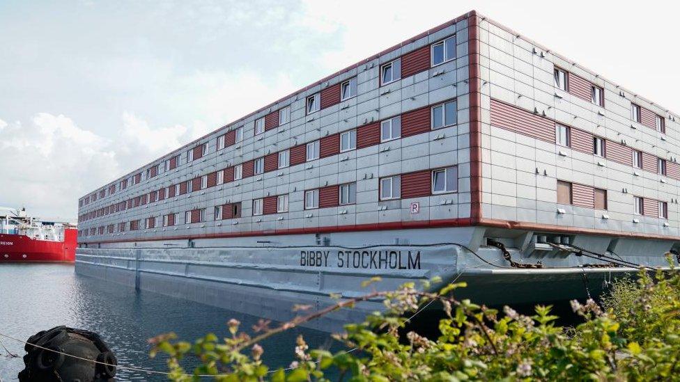 An exterior view of the Bibby Stockholm, the barge the UK government wants to use to house asylum seekers. It is three storeys high and has grey walls. It is berthed in Portland Port, and water is visible in the foreground of the image.