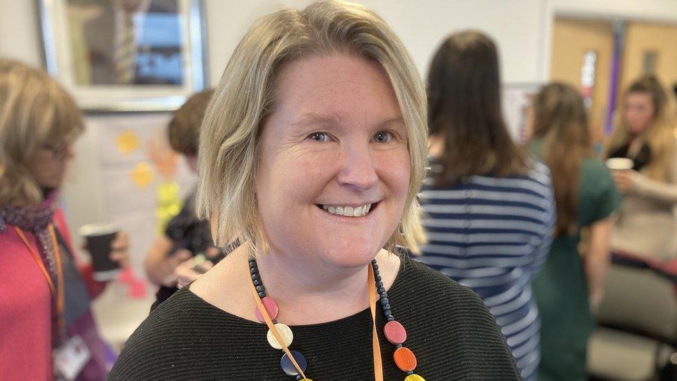 Emily Denne looks into the camera. She has short blonde hair and is wearing a dark stripy top and a big colourful necklace