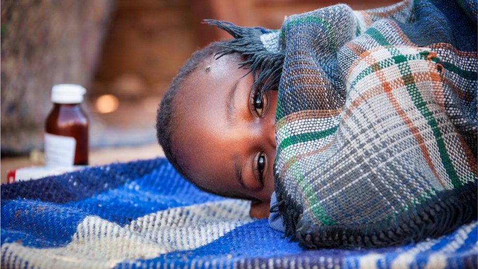 Toddler unwell with malaria, lying on a blanket with malaria medicine in the background