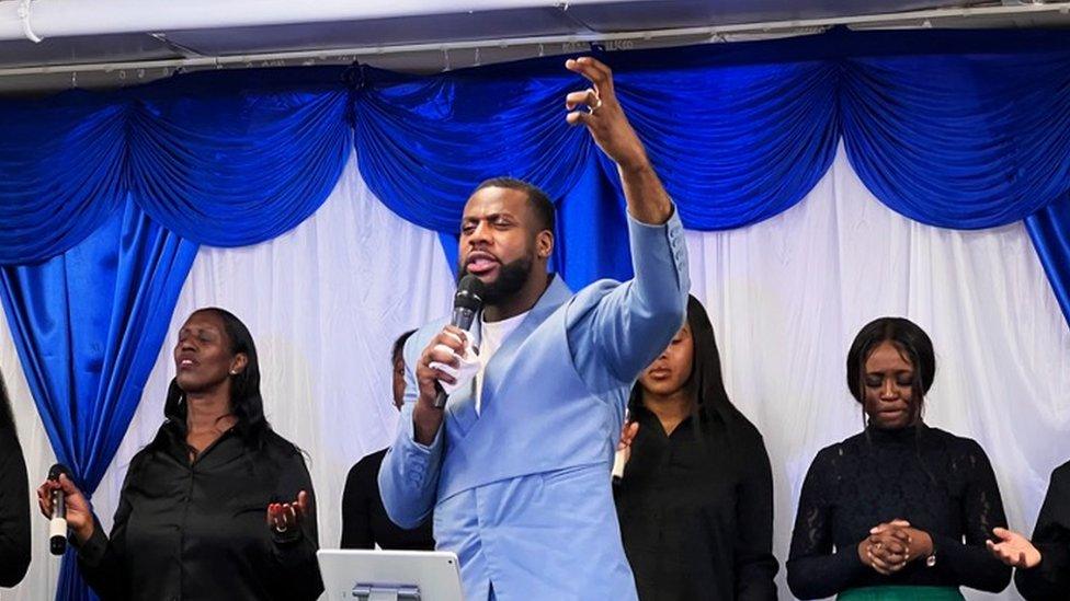 Pastor Onyeka standing on stage with a choir, speaking into a microphone with his eyes shut and hand raised