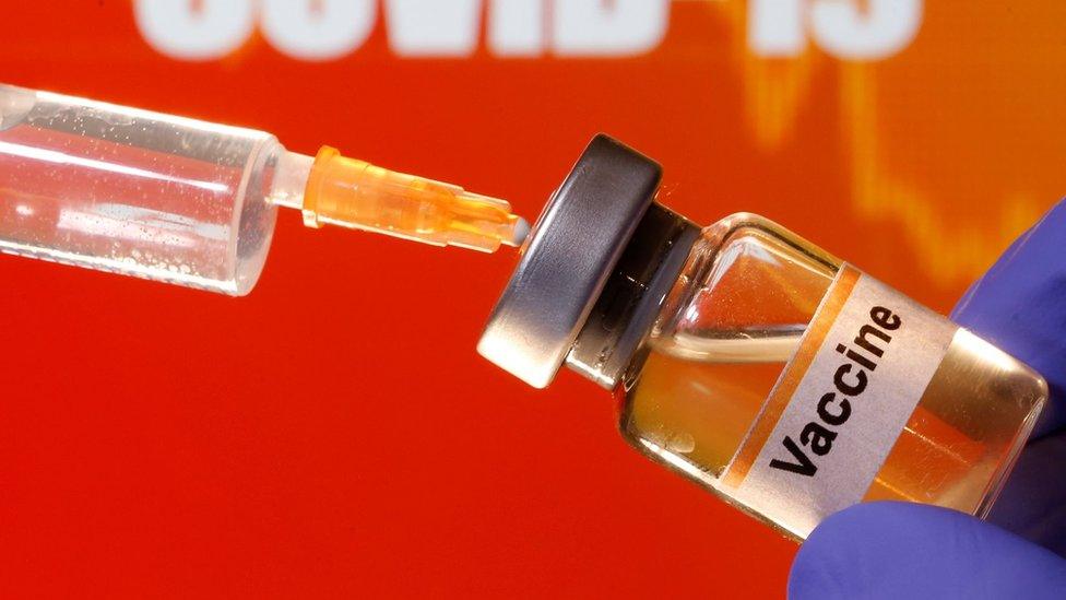 A small bottle labelled with a "Vaccine" sticker is held near a medical syringe