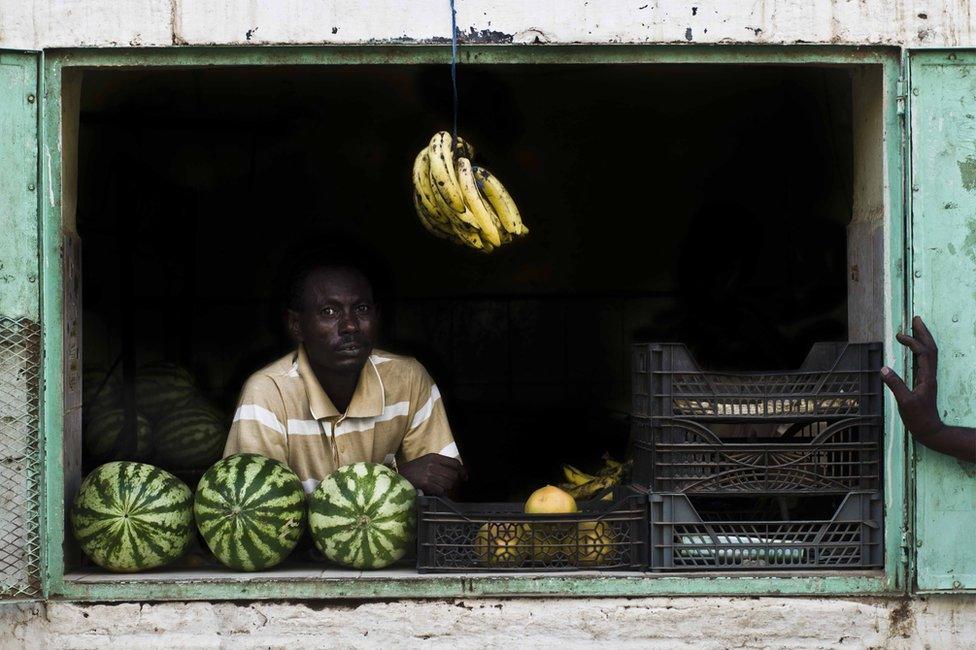 A fruit seller stands in front of his shop window with crates of fruit visible next and behind him