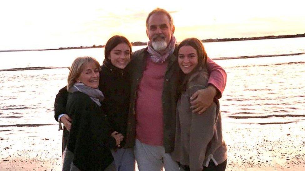 A mum and dad and two young men posing on a beach smiling at the camera