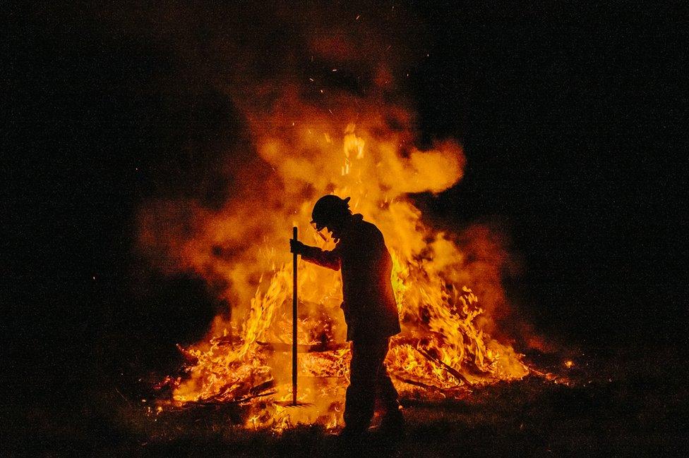 A firefighter, seen in silhouette, attends to a controlled fire with a rake