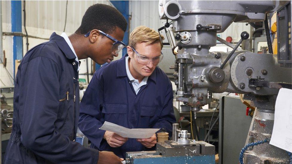 Engineering students in navy blue overalls and wearing safety goggles bend over a grey piece of machinery. One student has short, blond hair, the other, who is operating the machinery with his right hand, has short, black hair.