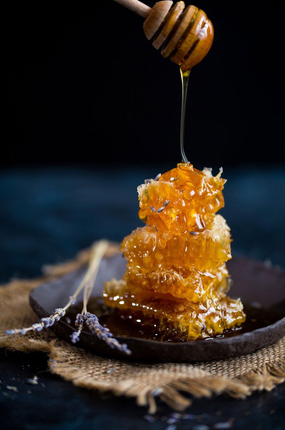 Runny Lavender Honey is being drizzled with a honey dipper over broken pieces of honeycomb decorated with dried lavender buds