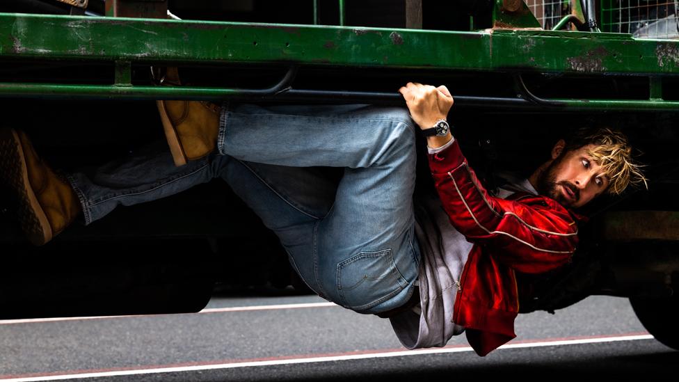 Ryan Gosling is Colt Seavers in The Fall Guy. Gosling is a white male actor in his early 30s. He has fair hair which is messy, a short brown beard and moustache and brown eyes. In the shot from the film, he hangs from the undercarriage of a truck. Ryan wears a red jacket, grey top, blue jeans and brown boots and has a concerned expression.