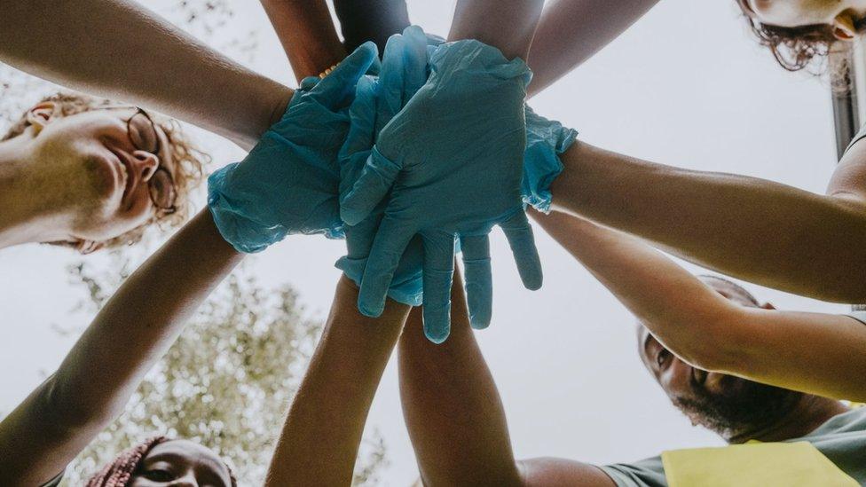 A group of people in high vis jackets with blue plastic gloved hands stand in a circle with their hands together in a show of teamwork