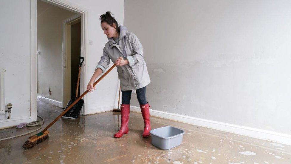 A woman is seen sweeping the floor of her home after flooding in Chesterfield. She is in wellington boots. A few inches of water are still on the floor.