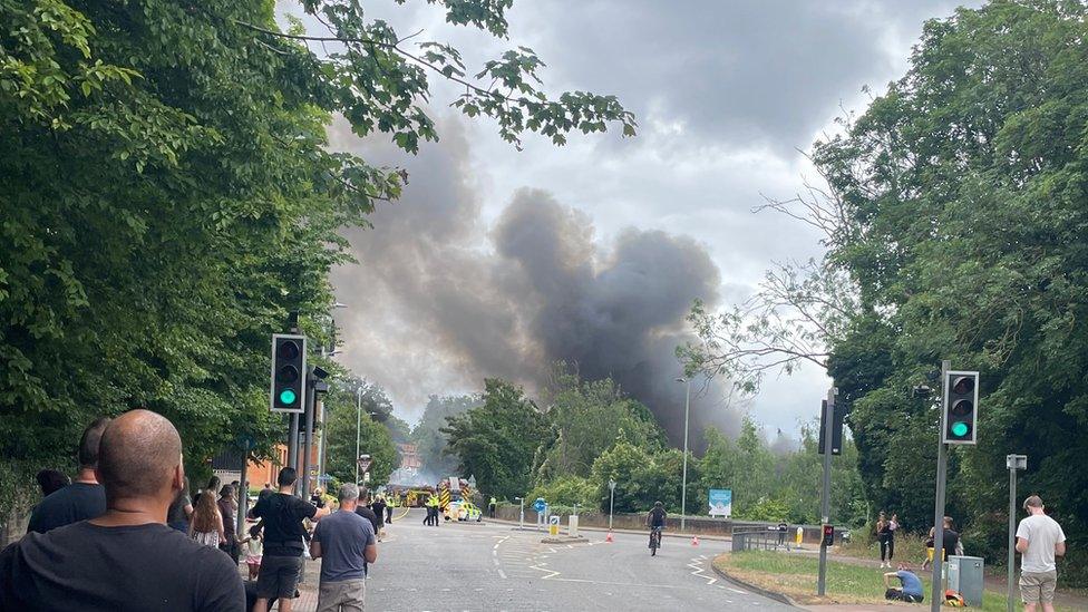 Plume of smoke above car workshop fire, with people looking on