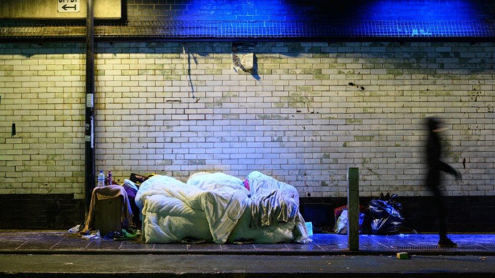 Commuters walk past three homeless people as they sleep in an exposed bed underneath a railway track