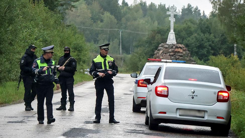 Police officers stand next to a car on a road with trees in the background, as they block the road following the incident