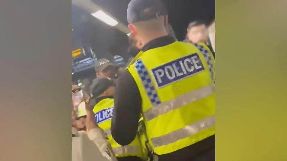 Greater Manchester Police officers are filmed in a crowd of people at Manchester Victoria. The backs of two officers wearing high-vis jackets and black caps can be seen, while one person films with a smartphone. 