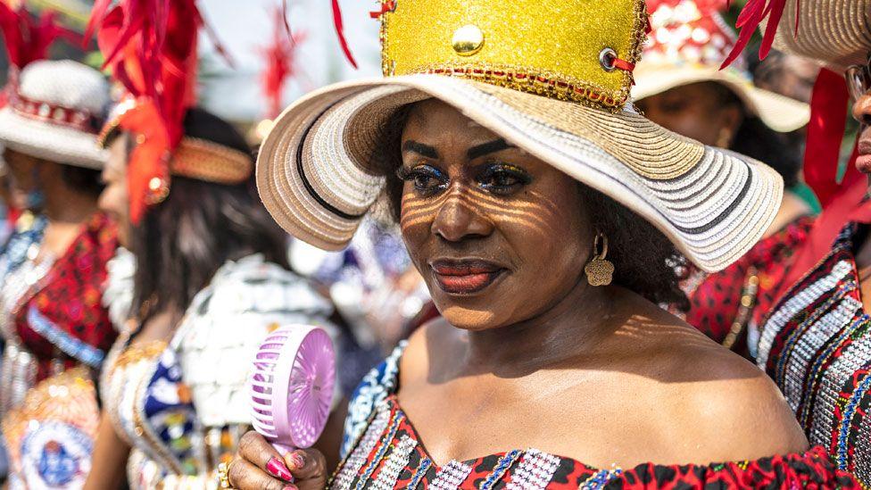 A woman in a large straw hat and off-the-shoulder outfit holds a pink electric fan as she takes part in the carnival in Calabar, Nigeria