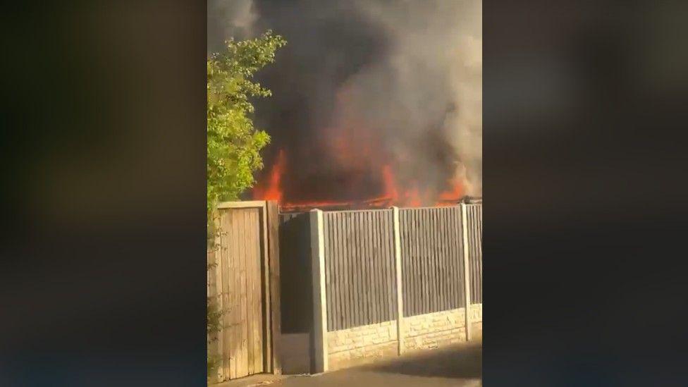 Smoke and orange flames can be seen above and behind a wooden fence