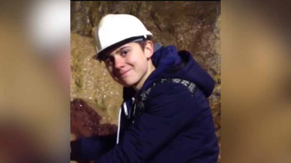 A teenage boy, wearing a white helmet and navy hoodie, smiles inside a cave.