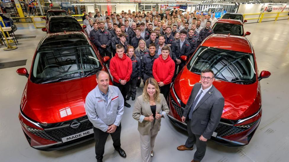 The inside of a factory with Kim McGuinness in the centre flanked by two men. There are six cars lined up with about a hundred workers between them.