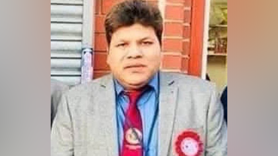 A man with brown hair and wearing a grey jacket, blue shirt, red tie and a red rosette.