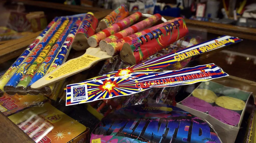 Sparklers, rockets and other fireworks sit on the counter of a shop, with the products featuring very colourful branding. 